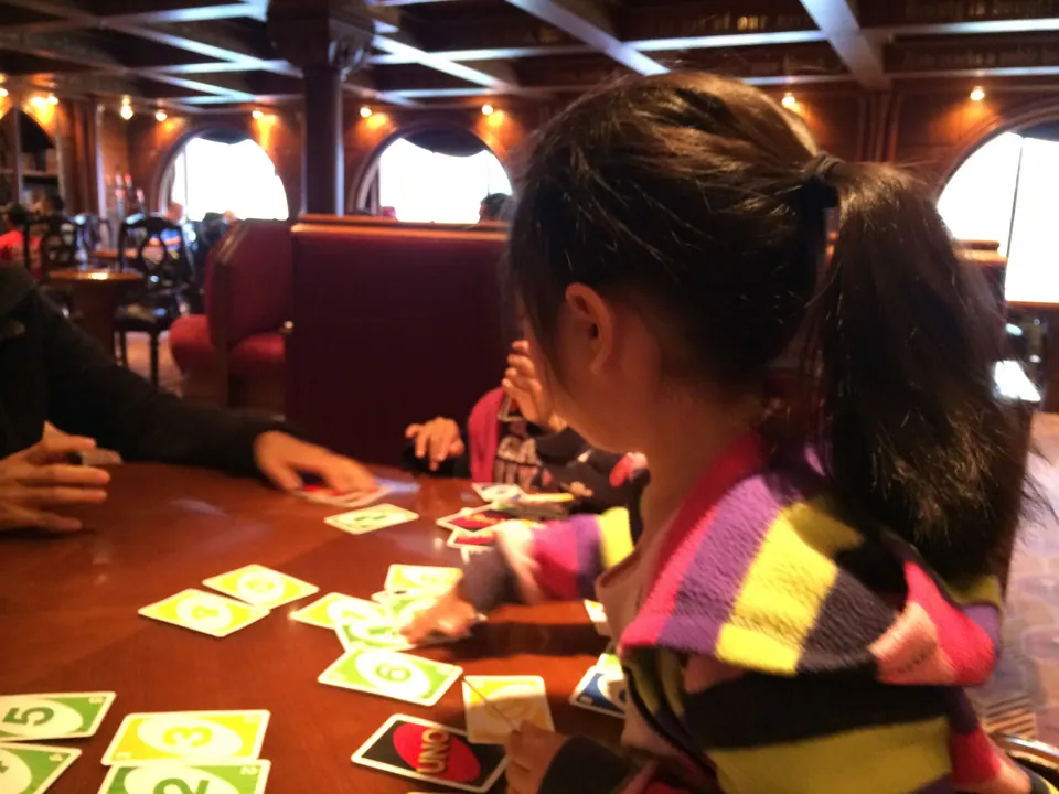 Green-backed UNO cards are spread over a polished wooden table in a lounge; a girl in a striped sweater plays a hand while another sits across from her.