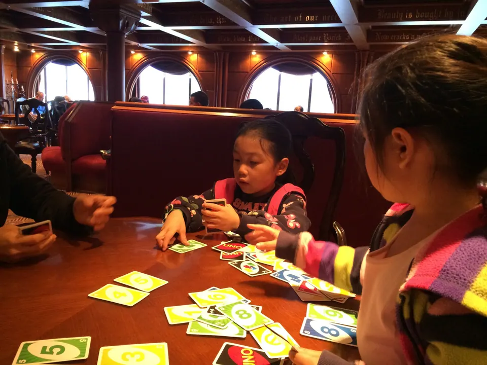 A girl in a navy jacket with a pink backpack strap studies her cards while a girl in a striped sweater leans in across a table strewn with UNO cards.