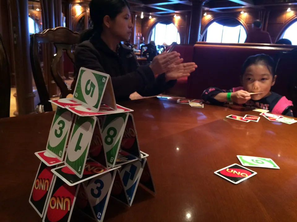 A tower of UNO cards stands on a polished wooden table in a warm-lit lounge, with a woman and a girl sitting behind it.