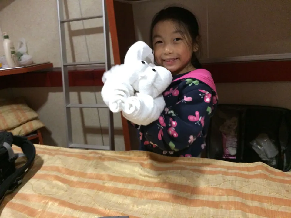 A smiling girl holds up an elephant folded from a white towel beside a bunk ladder in a small cabin.