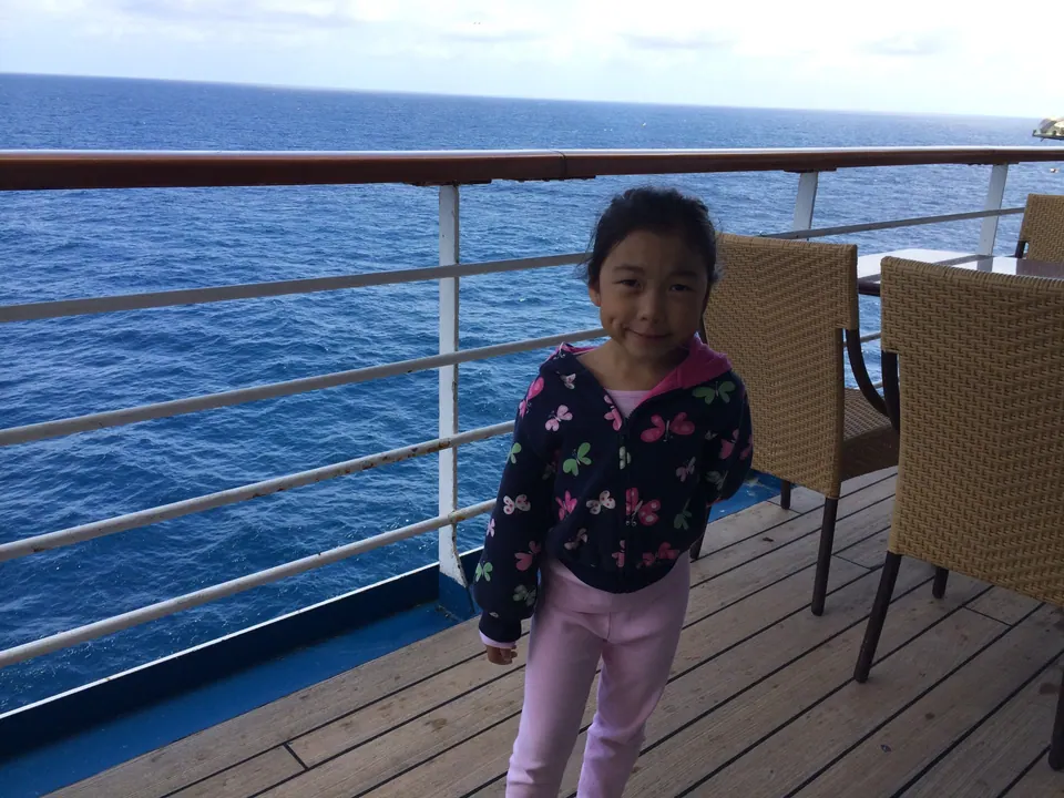 A girl in a butterfly-print jacket stands on a teak deck by a white railing, with open deep-blue ocean behind her.