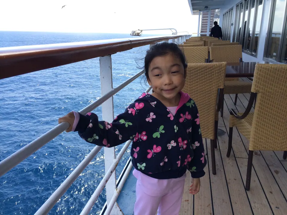 A girl in a butterfly-print jacket stands at the white railing of a teak deck with the blue Pacific behind her and wicker chairs on the far side.