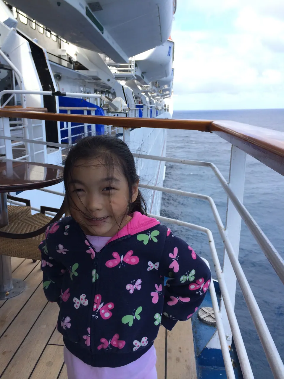 A girl in a butterfly-print jacket stands with her hands behind her back on a ship's deck, with railings and the sea behind her.