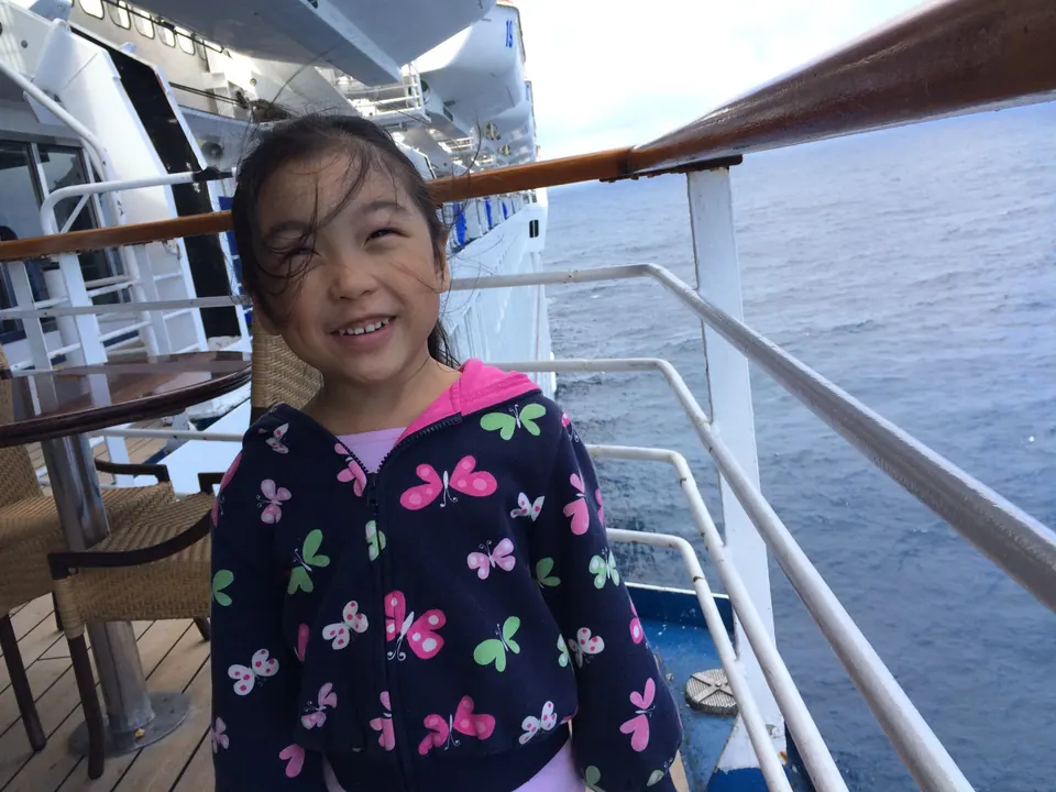 A grinning girl in a butterfly-print jacket with windblown hair on a ship's deck, the ship's white side and the ocean over her shoulder.