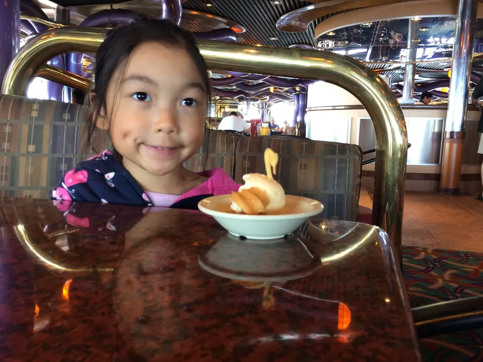 A smiling girl sits behind a bowl holding a swan-shaped dessert on a glossy table, with a brass rail and a bar area behind her.