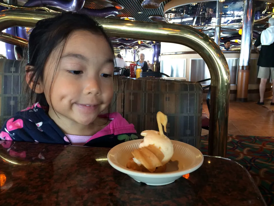 A girl smiles at a small bowl holding a dessert shaped like a swan, on a table with a brass rail in a bright lounge.