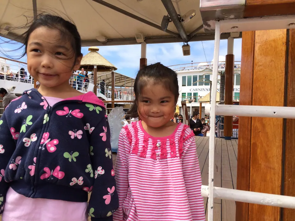 Two small girls stand under a canvas canopy on a sunny deck with an ice sculpture and other passengers behind them.