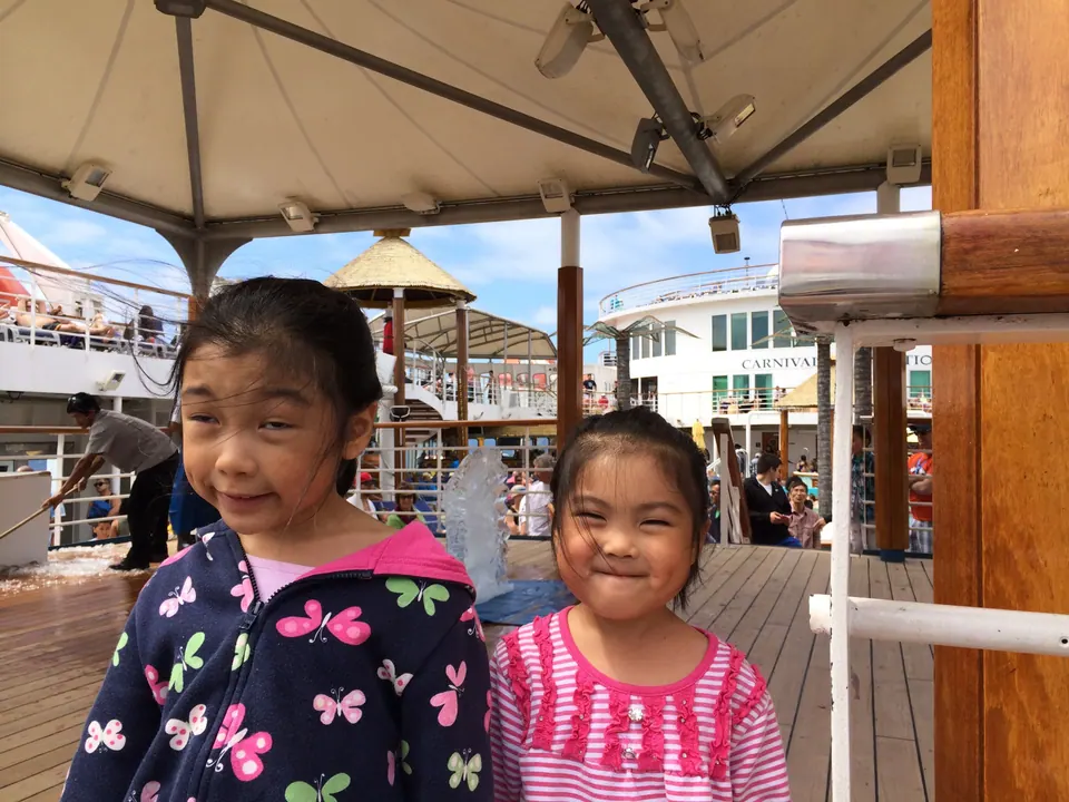 Two small girls stand under a white canopy on a sunny wooden deck; behind them a man works on a clear block of ice as passengers watch from the decks above.