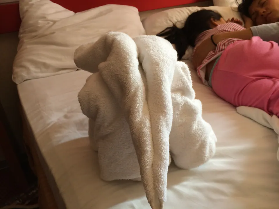 A towel animal is shaped on a bed while a small child in pink sleeps curled up beside it.
