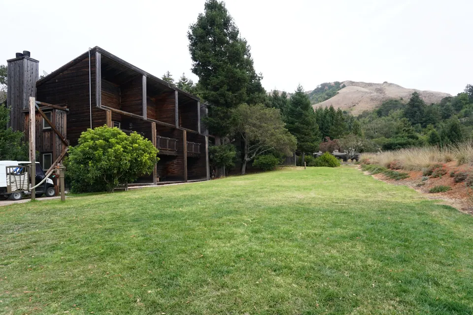 A dark wooden lodge building with balconies stands above a long green lawn, golden hills behind under a grey sky.