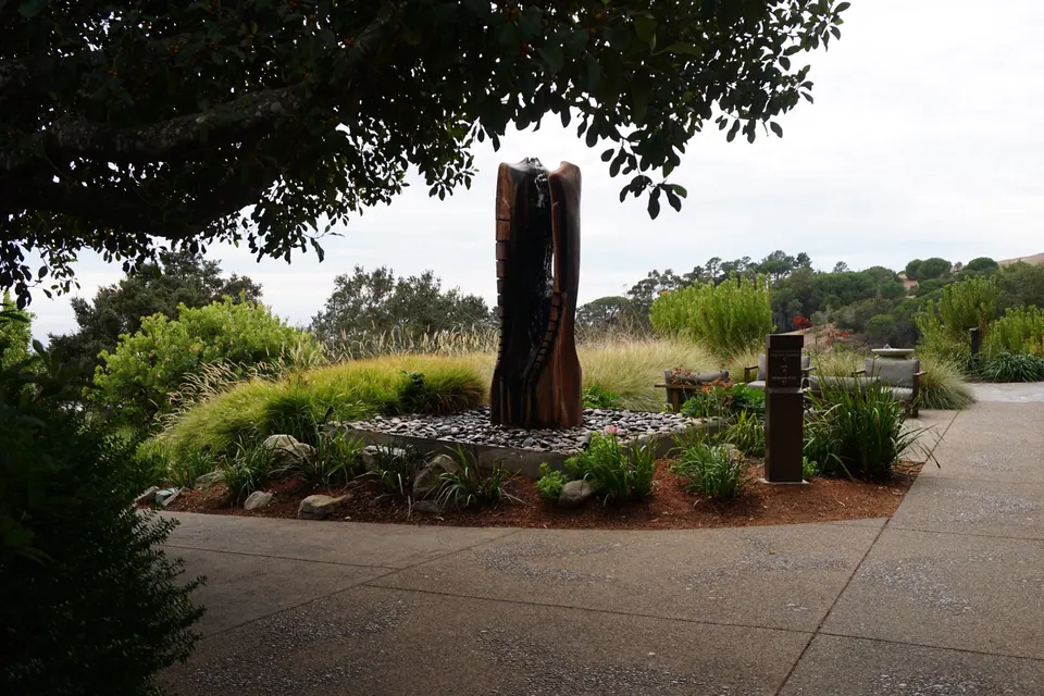 A tall carved wooden sculpture rises from a garden of grasses and shrubs at a bend in a wide path.