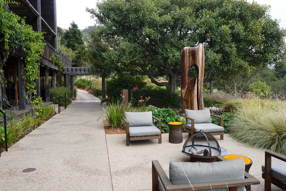 Gray lounge chairs and a metal fire bowl on a garden patio beside wooden lodge buildings, a carved sculpture behind.