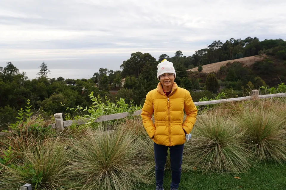 A woman in a yellow puffer jacket and white hat stands in front of clumps of grass and a rail fence, hills and sea behind.
