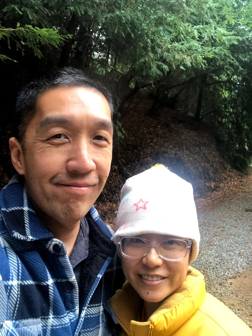 A man in a blue plaid shirt and a woman in a white knit hat and yellow jacket smile close together in a selfie on a gravel path.