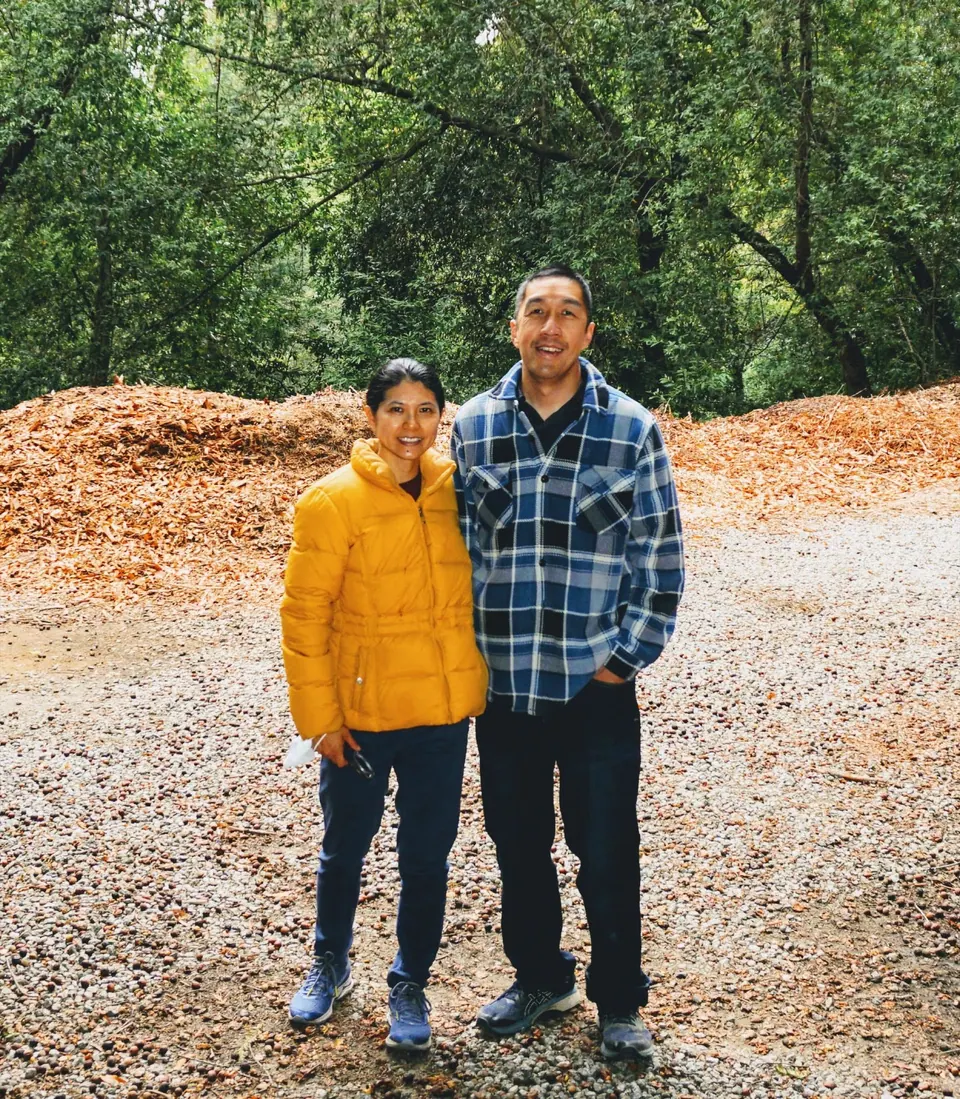 A woman in a yellow jacket and a man in a blue plaid shirt stand side by side in a clearing of woodchips among trees.