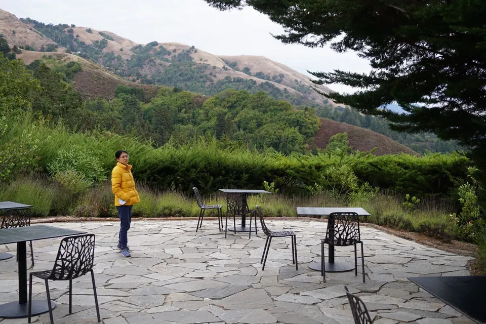 A woman in a yellow jacket stands on a flagstone terrace with empty tables and chairs, green hills behind.