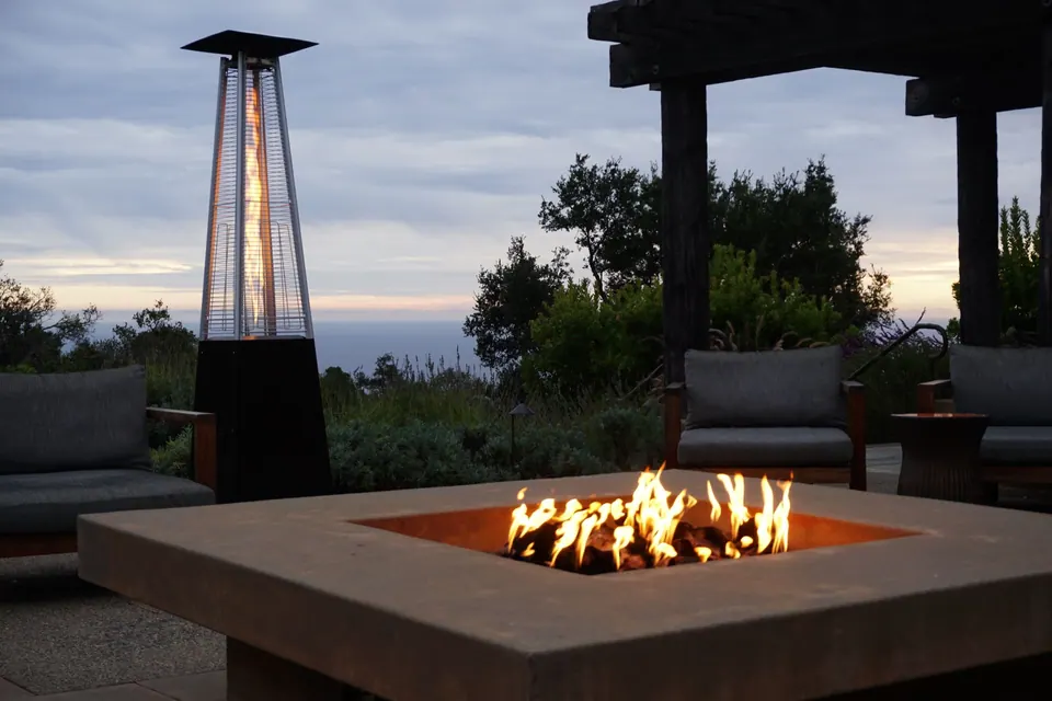 A gas fire pit burns on a patio at dusk, a patio heater and the sea faint behind.