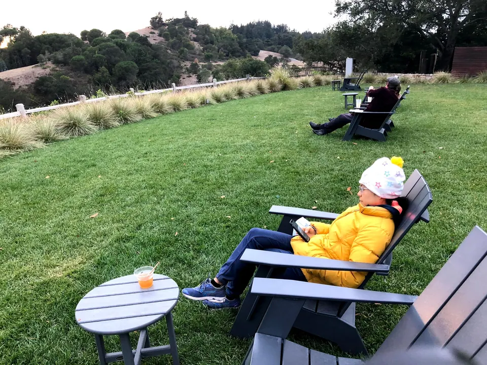 A woman in a yellow jacket and white hat reclines in a gray Adirondack chair on a green lawn, a drink on the table beside her.