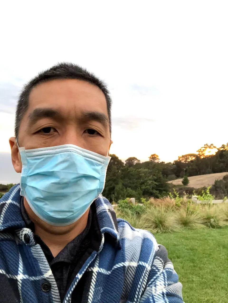 A man in a blue face mask and a blue plaid shirt takes a selfie against a pale evening sky.