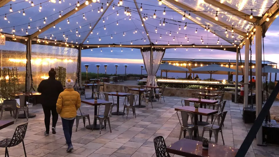 A woman in a yellow jacket walks across a stone terrace under a clear tent strung with lights, the sea glowing behind.