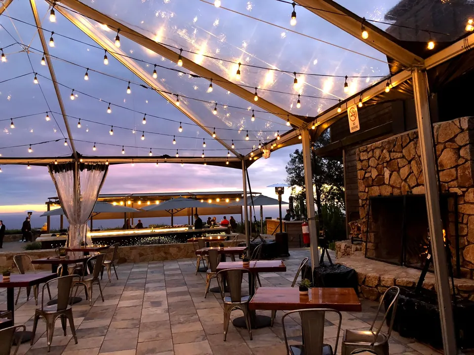 A clear tent hung with string lights over a stone terrace of small tables, with a stone fireplace and a pink sunset beyond.