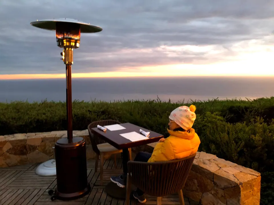 A woman in a yellow jacket and white hat sits at a table beside a patio heater, looking at an orange sunset over the sea.
