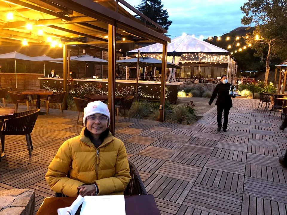 A woman in a yellow jacket and white hat smiles at a table on a wooden terrace, string lights and a staff member behind her.