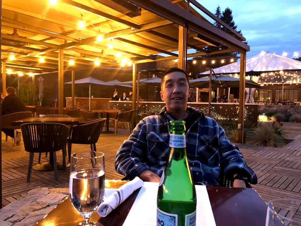 A man in a blue plaid jacket sits at a terrace table with a green bottle and a glass of water, string lights behind.
