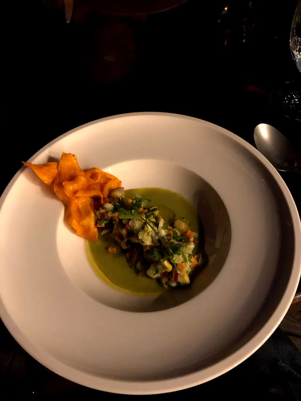 A white bowl holding a chopped salad on a green purée, topped with a curl of orange vegetable crisp.