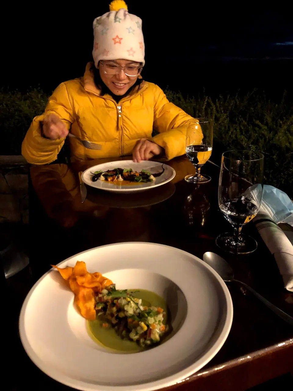 A woman in a yellow jacket and white hat eats at a table in the dark, two plates in front of her.
