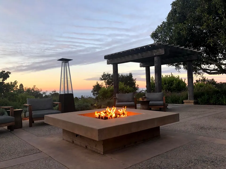 A gas fire pit burns on a patio beside a pergola and a patio heater, the sky turning orange over the sea.