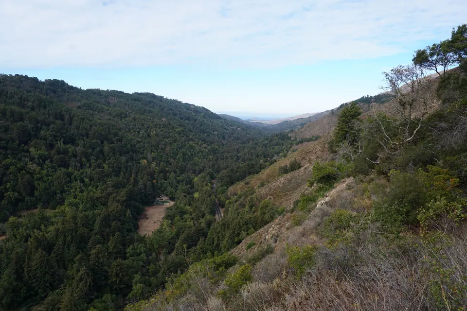 A deep, steep forested canyon opening to a hazy coastal plain under a pale sky.