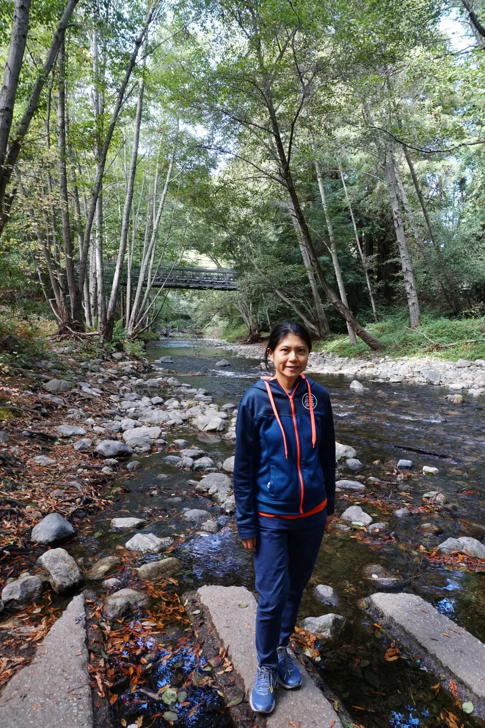 A woman in a blue jacket stands on concrete stepping stones in a rocky creek, the footbridge behind her.