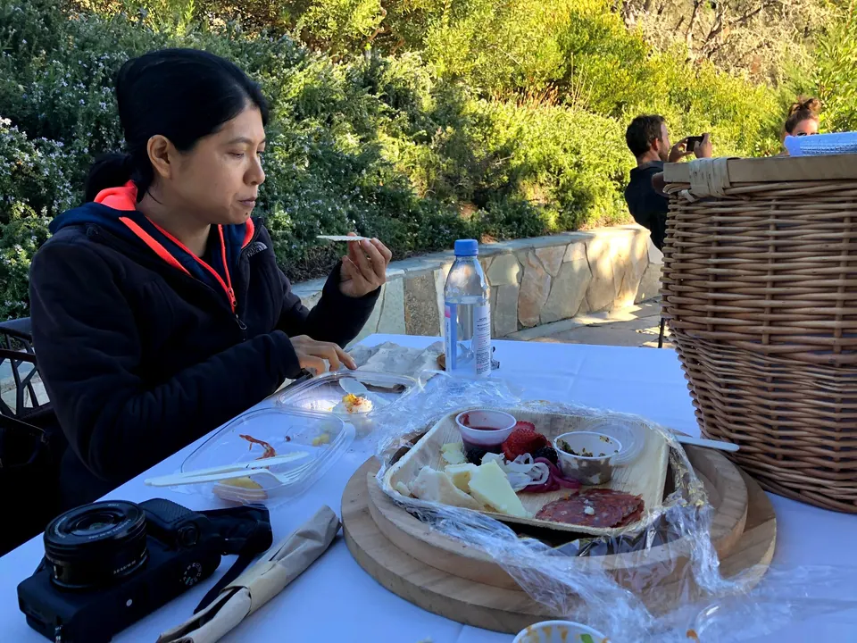 A woman in a dark jacket eats at a white-clothed table with a tray of cheese and cured meat, a camera and a wicker hamper beside her.