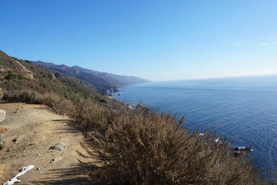 A dirt trail runs along a scrubby cliff above deep blue sea, with the Big Sur coast stretching away in haze.