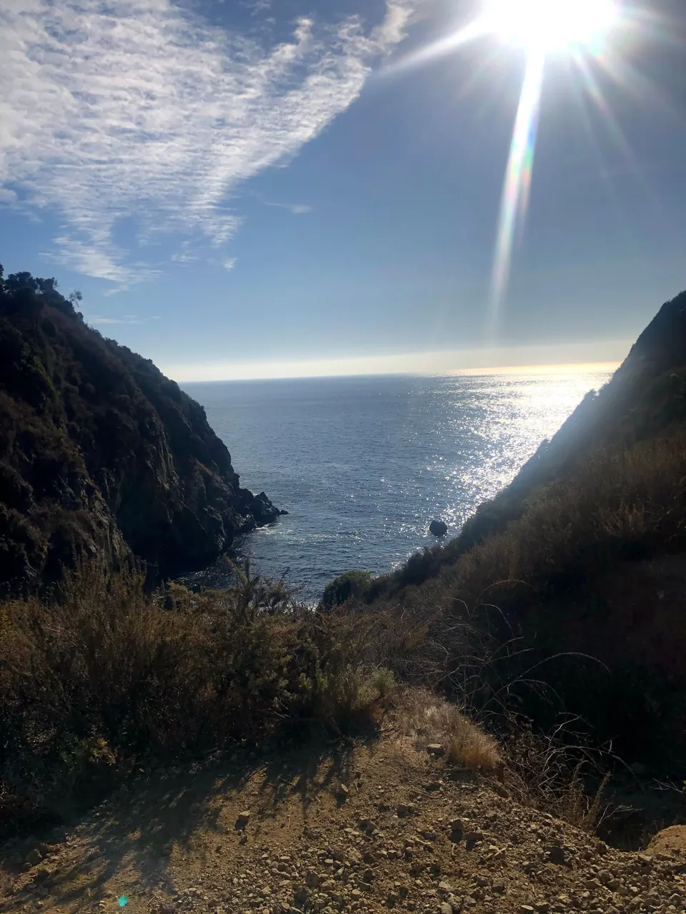 Late-afternoon sun sparkling on the sea between two steep, dark cliffs, seen from a trail.