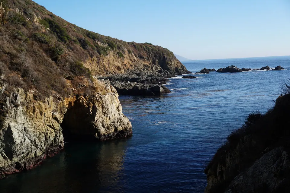 A rocky cove of deep blue water with a natural arch at the foot of a steep golden cliff.