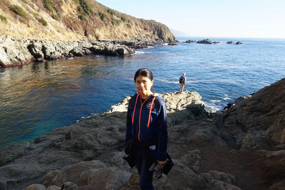 A woman in a dark jacket stands on a rock ledge over a blue cove, another hiker in the distance.