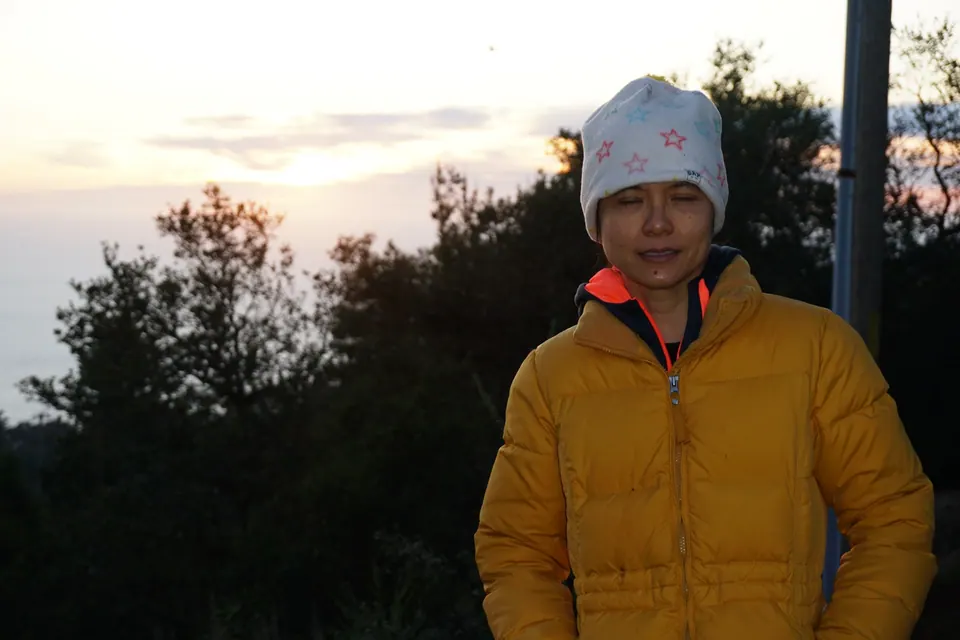 A woman in a yellow puffer jacket and white knit hat stands with her eyes closed in front of dark trees and a pale sky.