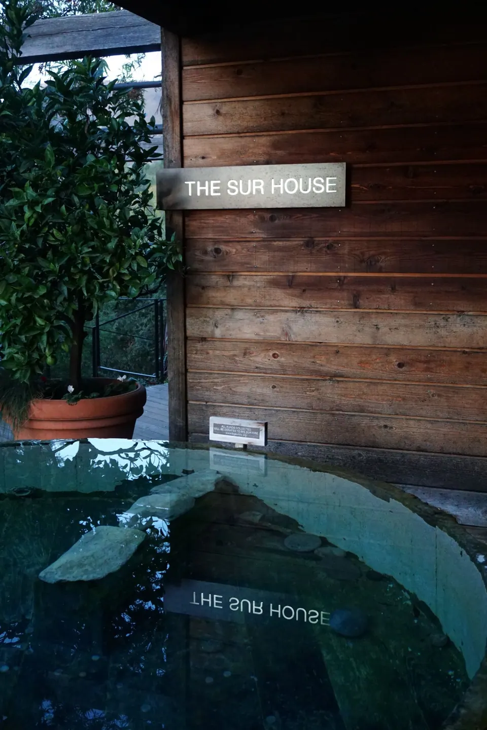 A wooden sign reading The Sur House on a shingled wall above a dark reflecting pool.