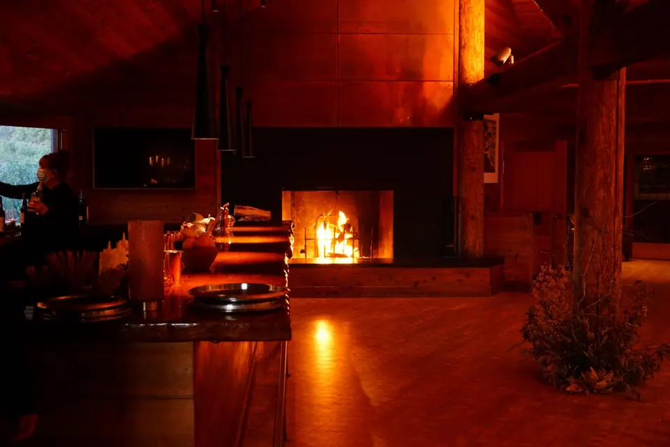 A stone fireplace blazes in a dark wooden dining room.