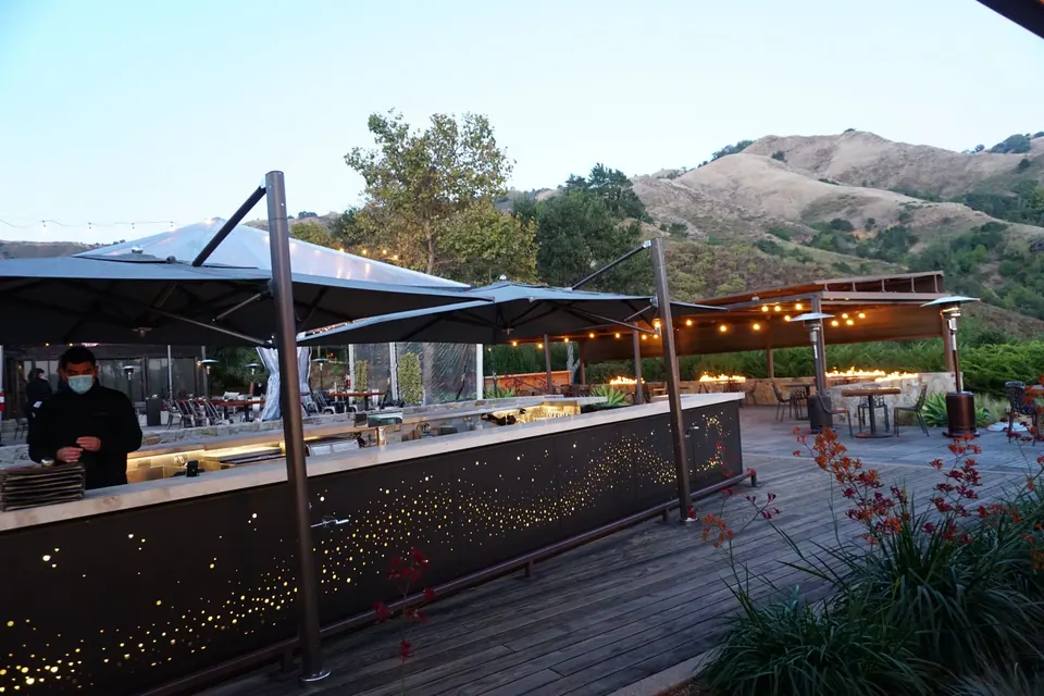 An outdoor bar under umbrellas with a screen of tiny lights, a masked server and green hills behind.