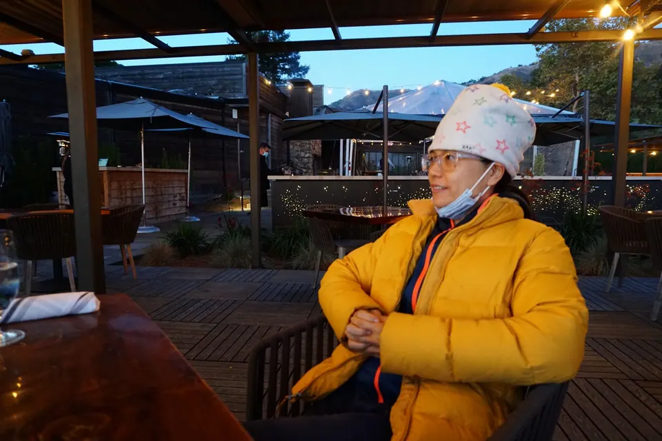 A woman in a yellow jacket and white hat sits at a terrace table, a face mask pulled below her chin.