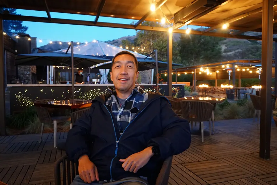 A man in a dark jacket sits at a table on a terrace under string lights, umbrellas and the hills behind.