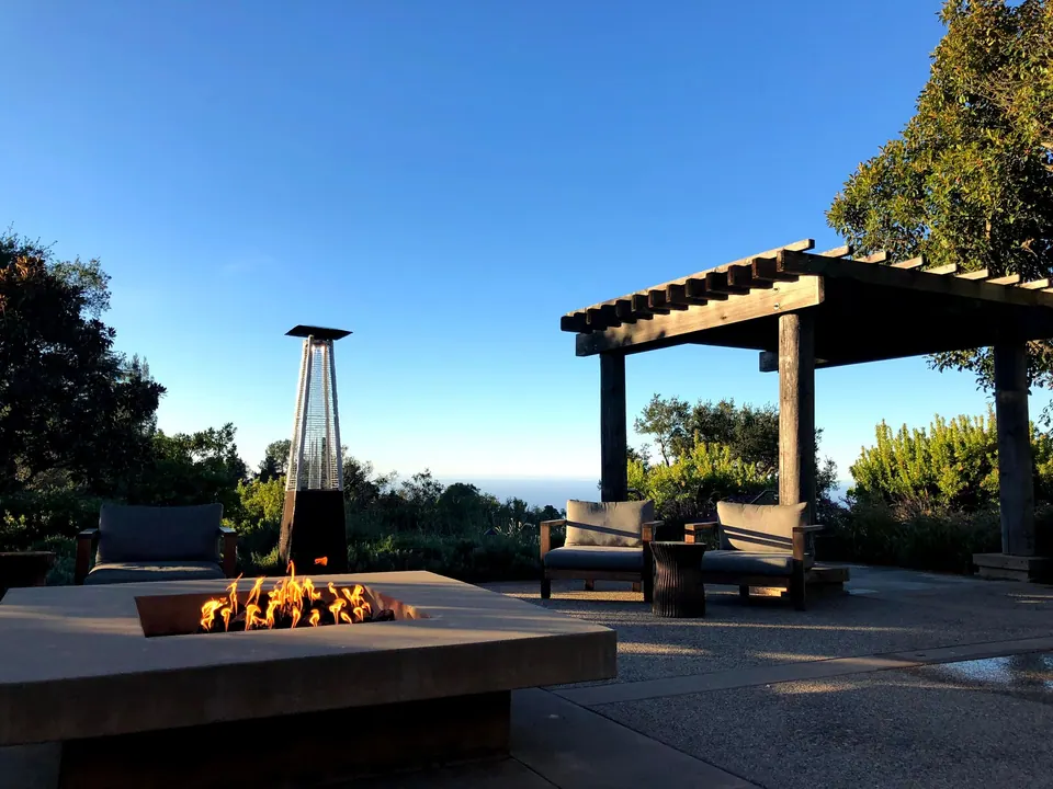 A gas fire bowl and pergola on a patio in low morning sun, a patio heater and the blue sea beyond.