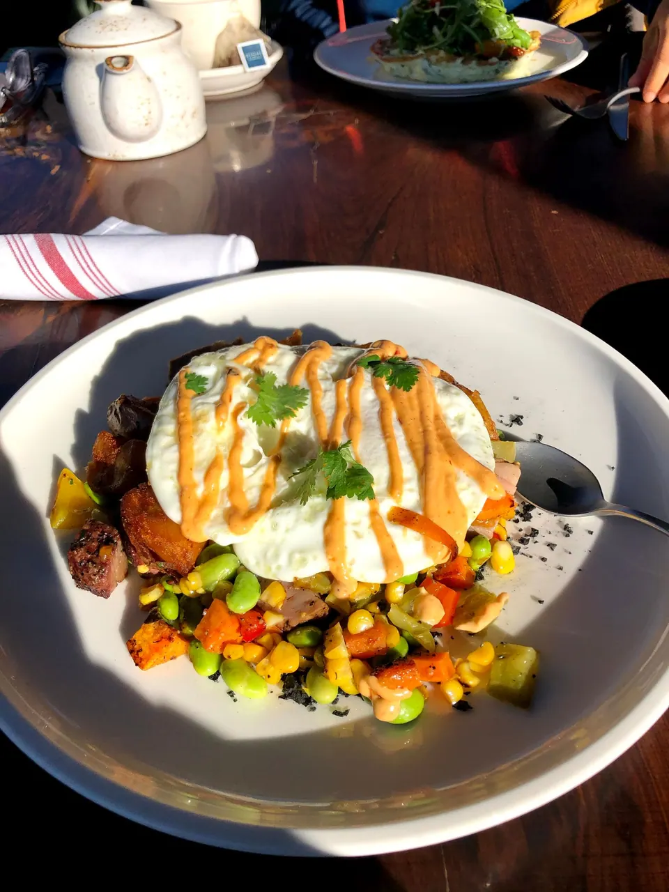 A white plate of a fried egg with chile drizzle over roasted vegetables and edamame on a sunny wooden table.