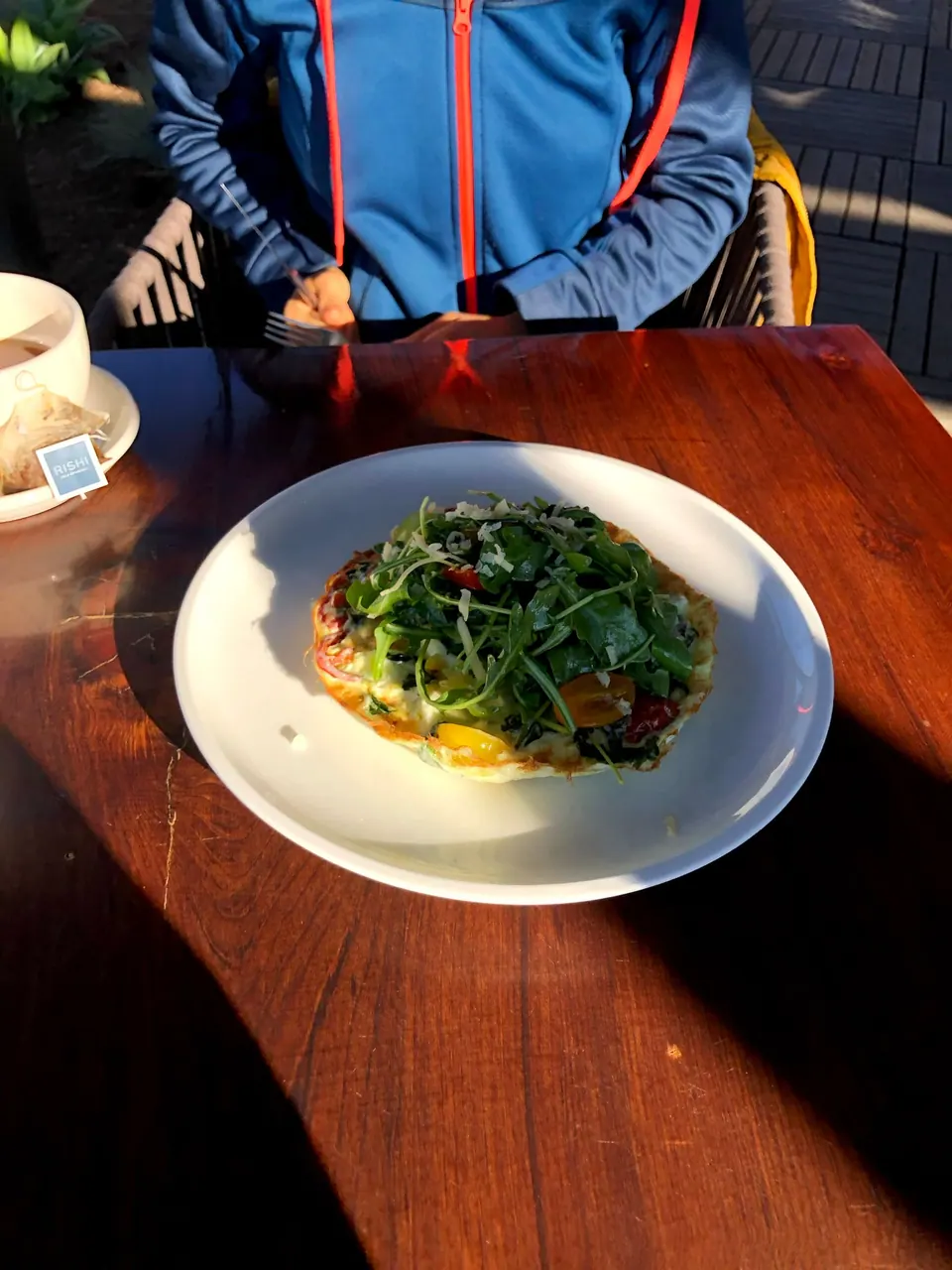 A white plate of arugula salad on a wooden table beside a teapot, a person in a blue jacket behind.