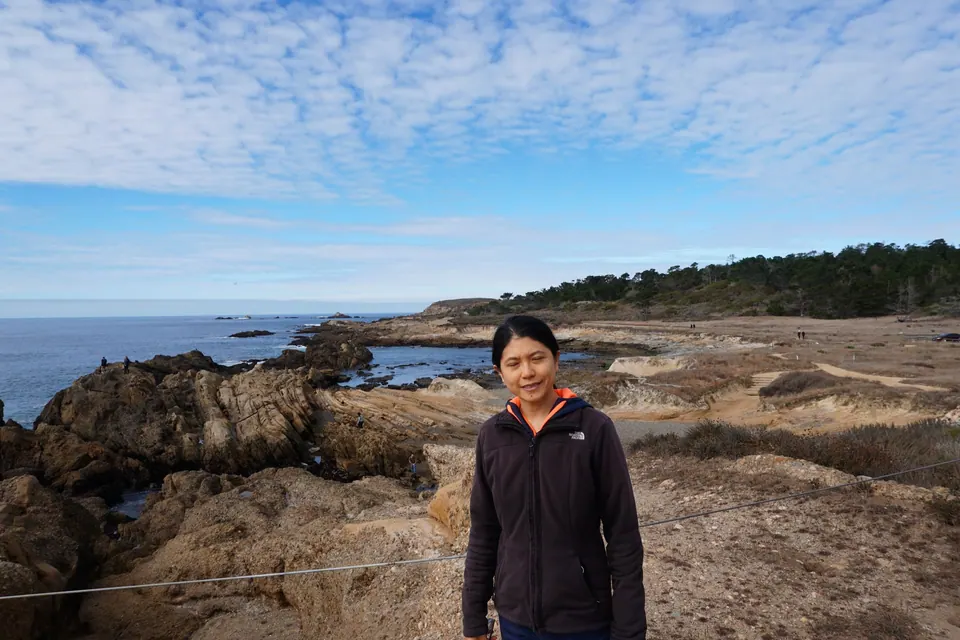 A woman in a black jacket stands on rocks with the coast behind her under mottled clouds.