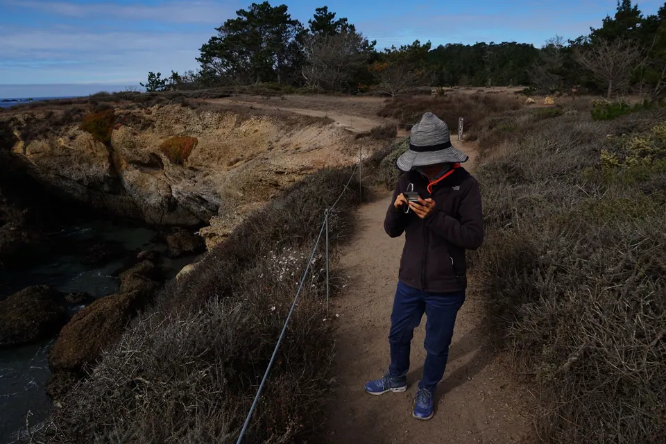 A woman in a sun hat looks at her phone on a coastal path, a cove and cliffs behind.