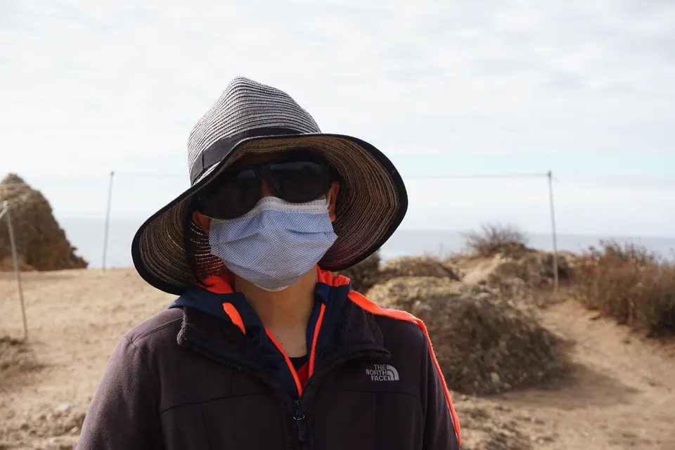 A woman in a sun hat, sunglasses and a face mask stands on a dry headland with the sea behind.